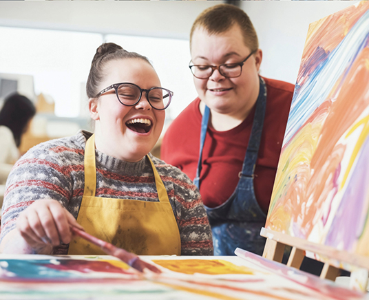 A female care worker engaging in the Community Support Program, painting artwork with a person who has intellectual and developmental disabilities (IDD).